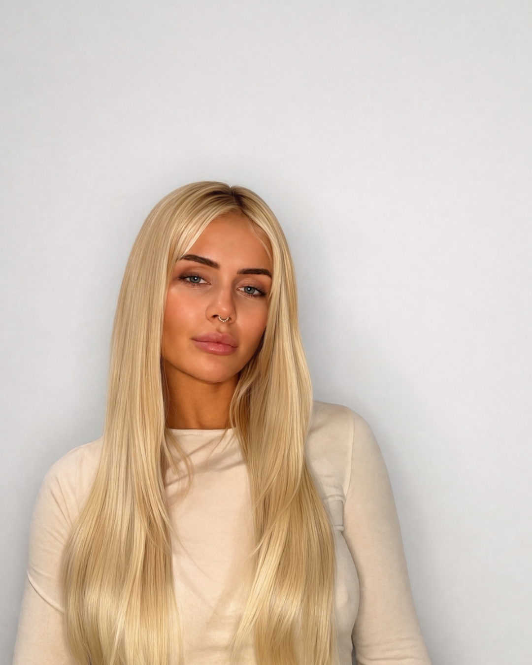 A woman with long, straight platinum blonde hair wearing a beige long-sleeve top, photographed against a plain light gray background.
